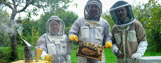 Three people in beekeeping suits holding a honeycomb frame outdoors.